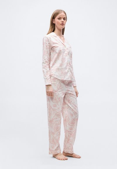 Νεαρή γυναίκα που στέκεται ξυπόλυτη, φορώντας ρόζ ανοιχτόχρωμο πουκάμισο με μακρύ μανίκι και φλοράλ σχέδιο, κουμπωτό, και χαλαρά παντελόνια που ταιριάζουν.