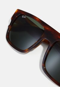 Ochelari de soare din material tartan, cu lentile întunecate, având rame groase și textură netedă. Logo-ul este vizibil pe tampla stângă.