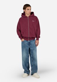 Young man wearing a maroon zip-up hoodie, loose-fitting blue jeans, and white sneakers standing with hands in pockets, looking to the side.