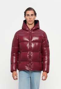 Man wearing shiny maroon puffer jacket with hood, standing against plain white background, hands relaxed by sides.