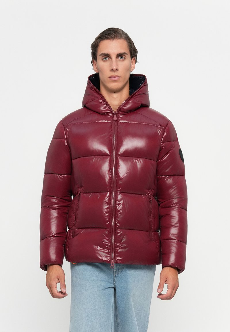 Man wearing shiny maroon puffer jacket with hood, standing against plain white background, hands relaxed by sides.