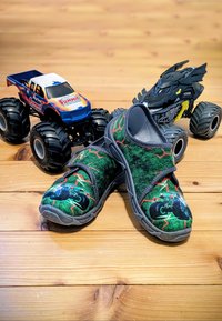 Groene stoffen instap schoenen met zwarte en neon monstertruck ontwerpen, met grijze accenten en een stevige rubberen zool. Twee speelgoed monstertrucks in de buurt.