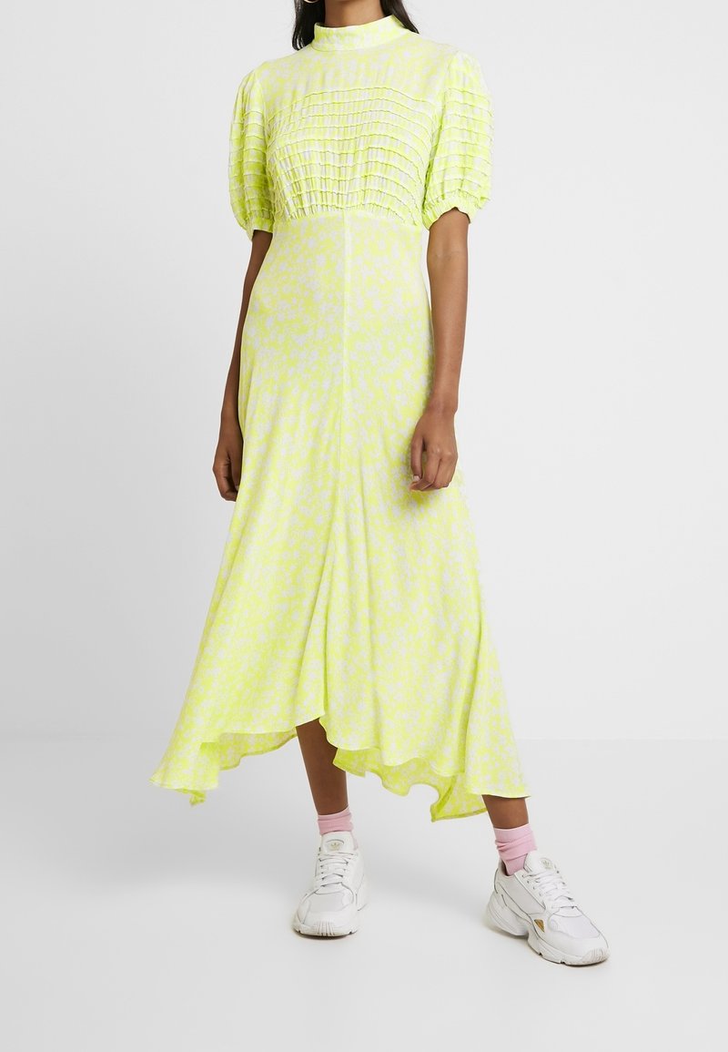 Robe jaune pâle à fleurs avec manches bouffantes, corsage froncé, ourlet asymétrique, tissu léger. Portée avec des baskets blanches et des chaussettes roses.