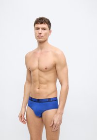 Άνδρας χωρίς μπλούζα, στέκεται μπροστά σε λευκό φόντο, φοράει μπλε σλιπ Puma με μαύρη μέση, κοιτάζει μπροστά με ουδέτερη έκφραση.