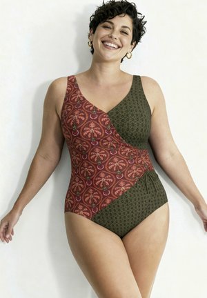 Glunderende vrouw met kort krullend haar draagt een mouwloze, patroonbadpak die rode en groene delen combineert, staand tegen een effen muur.