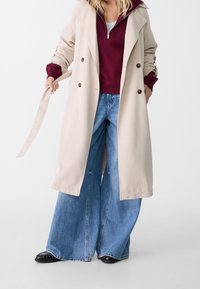 Beżowy płaszcz trench z burgundowym hoodie, w połączeniu z szerokimi niebieskimi dżinsami. Posiada guziki, pasek oraz czarne buty.