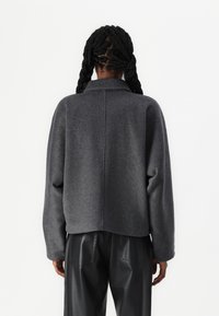 Veste en mélange de laine grise avec une coupe décontractée, une texture épaisse et un col. Le dos présente un détail de couture. Associée à un pantalon en cuir noir.