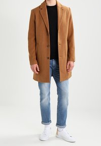 Brun ullblandad ytterrock med spetsiga slag, två knappar och sidofickor. Kombinerad med en svart t-shirt, blå jeans och vita sneakers.