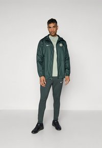 Herrarnas Nike sportkläder med en mörkgrön jacka, ljusgrön skjorta och matchande figursydda byxor. Materialen ser släta ut med logotypdetaljer.