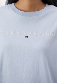 Světle modré bavlněné tričko s vyšitým nápisem "TOMMY JEANS" bílou barvou, pod kterým se nachází malá vlajková nášivka v červené, bílé a modré barvě.