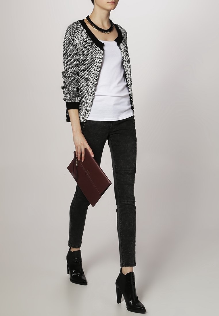 Femme portant un cardigan à motifs noir et blanc, un top blanc, un jean skinny noir, des bottines noires, tenant une pochette bordeaux.