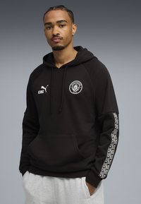 Ung mand med fletninger iført en sort Manchester City hoodie med hvide logoer og grå sweatpants, stående foran en grå baggrund.