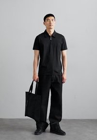 Black polo shirt with a zip collar, paired with loose-fitting black trousers and a flat black tote bag. Items are made from smooth fabric.