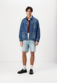 Veste en denim bleue, avec poches plaquées et boutons. Chemise blanche en dessous, accompagnée de shorts bleu clair et de chaussures noires à enfiler.