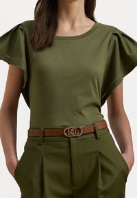 Vihreä oliivinvärinen t-paita, jossa on vaakasuunnassa laskostetut lyhyet hihat, yhdistettynä korkeavyötäröisiin khaki-housuihin ja ruskeaan vyöhön, jossa on kultainen solki.