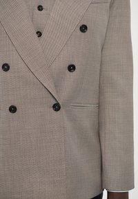 Kétgombos zakó világos bézs textúrált anyagból, fekete gombokkal, klasszikus hajtókákkal és oldalsó zsebekkel.