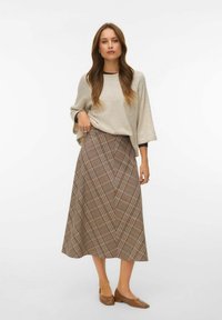 Femme aux longs cheveux portant un haut beige ample, une jupe midi à carreaux marron et des chaussures marron à talons bas, se tenant devant un fond blanc.