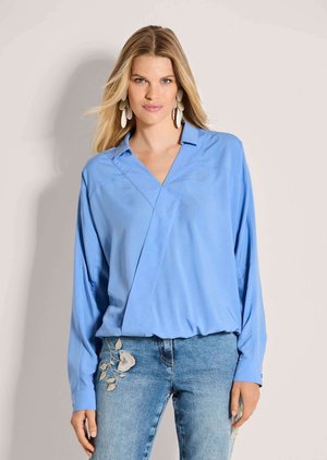 Femme blonde portant un chemisier bleu oversize à col en V et un jean bleu clair brodé d’un motif floral, debout devant un fond uni.