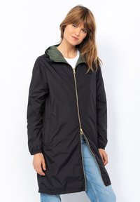 Donna in un lungo impermeabile nero con cappuccio e zip dorata, foderato di verde, camicia bianca e jeans azzurri chiari, in piedi su uno sfondo bianco.