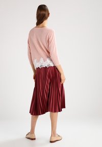 Maglione rosa chiaro con dettagli in pizzo all'orlo, abbinato a una gonna plissettata borgogna. Tessuto morbido, maniche a tre quarti e scarpe basse.