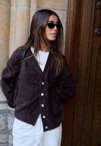 Mörk, fluffig cardigan med V-ringning och fem knappstängningar, i kombination med en vit topp och ljusa byxor. Svarta solglasögon används.