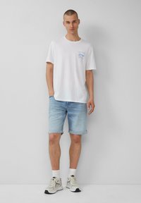 T-shirt en coton blanc avec un graphique bleu, short en denim clair avec ourlets retroussés et baskets claires. Tenue simple et décontractée sur un fond neutre.