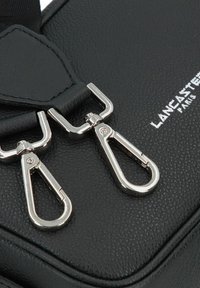 Dettaglio di una borsa in pelle nera con texture, dotata di chiusure e cuciture in argento. Il logo del marchio è impresso in bianco.
