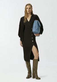 Vestido negro de canalé con escote en V profundo y abertura lateral, con botones plateados. La modelo lleva un bolso acolchado de color azul claro y botines verdes hasta la rodilla.