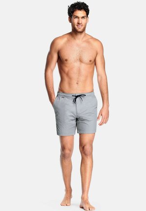 Man met krullend donker haar, gekleed in lichtgrijze zwemshorts met een zwarte trekkoord, staat blootsvoets tegen een effen witte achtergrond.