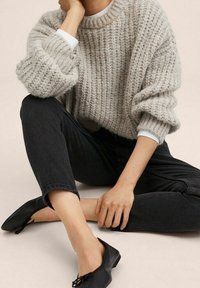 Un suéter de punto grueso y gris claro con textura de canalé, combinado con jeans negros y zapatos planos negros, sentado en una pose casual.