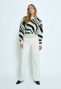 Maglione a righe bianco e nero, con texture e collo alto, abbinato a pantaloni bianchi a vita alta e gamba larga, e scarpe nere.