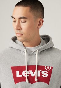 Grijze hoodie van katoen, met een groot rood Levi's logo. Het heeft witte trekkoorden, een buidelzak en een zachte textuur.