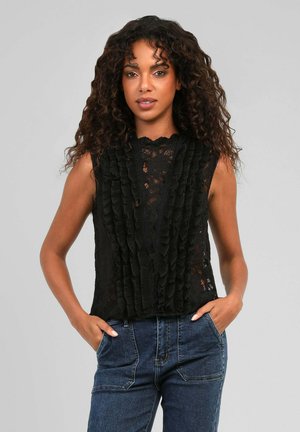 Femme aux cheveux bouclés portant un top en dentelle noire sans manches avec des volants et un jean bleu avec les mains dans les poches.