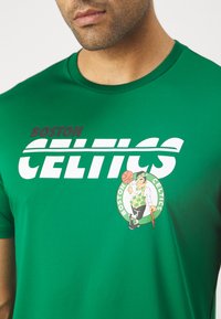 Zöld, rövid ujjú póló, amelyen a "BOSTON CELTICS" felirat található vastag fehér betűkkel, valamint egy logo, amelyen egy kobold látható kosárlabdával.