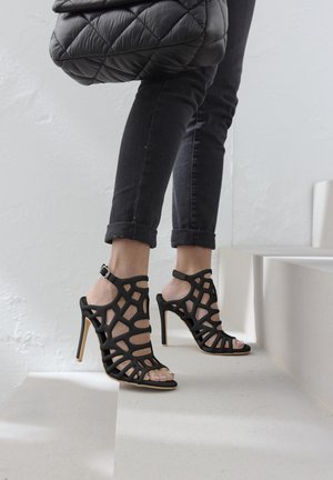 Black high-heeled sandals with a geometric cut-out design, open toe, and ankle strap. Worn with rolled-up dark jeans.
