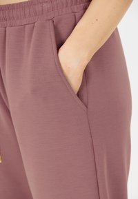 Mauve sportieve broek met een elastische tailleband en zijzakken, gemaakt van een zachte, gestructureerde stof. Gouden accenten zijn aanwezig.