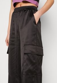 Gros plan d'une personne portant un pantalon cargo large en satin noir avec une ceinture élastique et une main dans la poche, assorti d'un pull court violet.