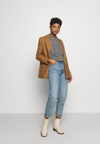 Blazer castanho sobre uma blusa azul floral, jeans claros de cintura alta e botas de tornozelo creme com um acabamento texturizado.