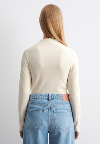 Gerippter, cremefarbener Langarm-Oberteil mit gewelltem Ausschnitt, kombiniert mit hoch taillierten, hellblauen Jeans mit zwei Gesäßtaschen.