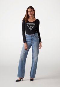 Musta pitkähihaisena paita, jossa on kolmiomainen logonmuotoilu, yhdistettynä korkeavyötäröisiin vaaleansinihin farkkuihin ja mustiin kärkitossuihin.