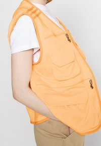 Orange ärmlös väst med flera fickor, dragkedjedetaljer och axelränder, bärs över en vit t-shirt och visar en slät tygtextur.