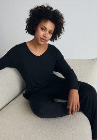 Svart långärmad topp med rundad halsringning, matchad med svarta byxor, tillverkade av ett mjukt, stretchigt tyg. Modellen sitter på en ljus soffa.