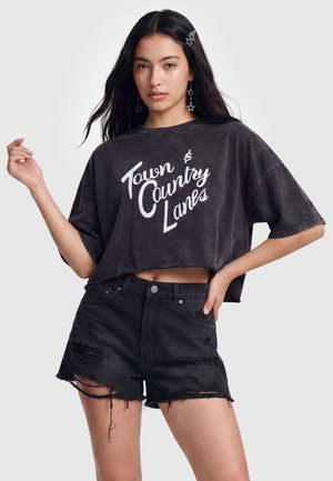 Ung kvinde poserende i sort cropped T-shirt med "Town & Country Lanes" tekst og sorte slidte denimshorts mod en hvid baggrund.
