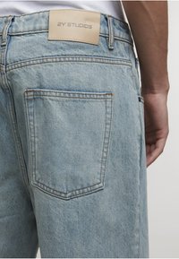 Achteraanzicht van lichtblauwe spijkerbroek met een gestikte achterzak en een beige label met de tekst "2Y STUDIOS" op de tailleband.