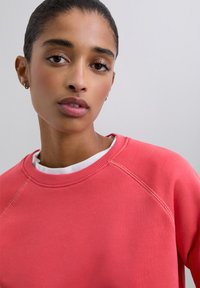 Sweat-shirt corail avec un col rond et des manches raglan. Texture lisse avec des détails de couture, superposé à un col de chemise blanc.