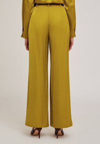 Breda byxor i senapsgul, tillverkade av ett mjukt tyg. Har en bälte i midjan och en lätt lyster, som matchas med bruna högklackade skor.