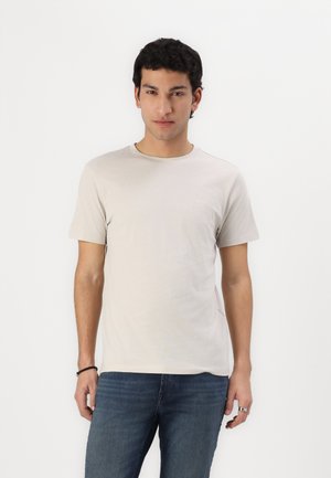 Hellbeige Baumwoll-T-Shirt mit Rundhalsausschnitt; kurze Ärmel; zeigt ein dezentes Logo auf der Brust; kombiniert mit blauen Jeans.