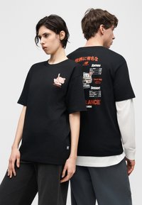 Twee modellen dragen zwarte New Balance t-shirts met een ramenkomgrafiek op de voorkant en een ramenmenutekst met Japanse karakters op de achterkant.