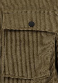 Olivgrüner Cordstoff mit vertikalen Rillen, ausgestattet mit einer großen Tasche mit einem schwarzen Druckknopfverschluss auf der linken Seite.