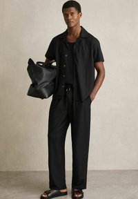 Camisă scurtă cu mânecă, neagră, cu dungi, cu nasturi, pantaloni lungi asortați și sandale; modelul ține o geantă mare de umăr neagră. Textură netedă.